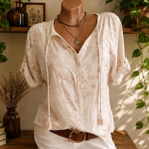Puede incluir: Una blusa de color crema con un sutil estampado floral marrón claro, escote en V con largos lazos trenzados y mangas cortas con trabillas abotonadas. La prenda se combina con un cinturón de cuero marrón y un collar de cuentas en capas.