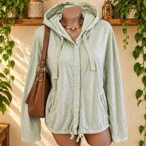 Puede incluir: Una chaqueta con capucha de manga larga en color verde salvia claro, con cierre frontal de botones, capucha con cordón y dos bolsillos laterales. Un bolso de hombro de cuero marrón con detalle de borla cuelga sobre el hombro de la prenda.