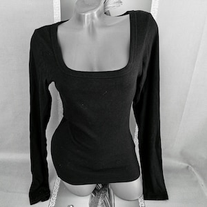 Könnte beinhalten: Schwarzes Langarm-Top mit quadratischem Ausschnitt. Das Oberteil ist aus geripptem Strickstoff gefertigt und hat eine figurbetonte Silhouette. Die Ärmel sind lang und enden an den Handgelenken. Das Oberteil wird auf einer Schaufensterpuppe präsentiert.