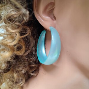 Può includere: Primo piano di un orecchio con un grande orecchino a cerchio azzurro. L'orecchino ha una lucentezza perlescente e un design liscio e curvo. I capelli biondi ricci della persona sono visibili sullo sfondo.
