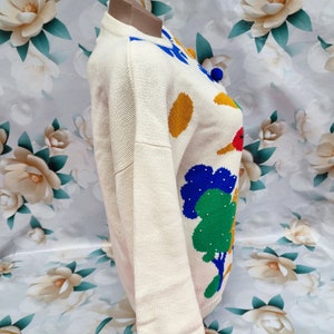 May include: A cream-coloured long-sleeve knit jumper featuring a colourful graphic pattern of green trees, yellow circles, and blue accents. The jumper has a round neckline and a ribbed hem, showcasing a textured knit construction throughout the garment.