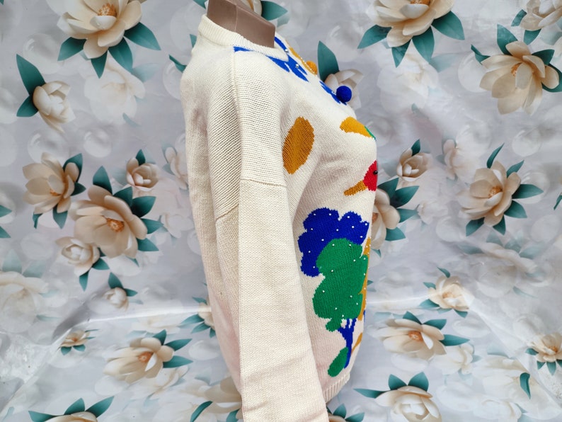 May include: A cream-coloured long-sleeve knit jumper featuring a colourful graphic pattern of green trees, yellow circles, and blue accents. The jumper has a round neckline and a ribbed hem, showcasing a textured knit construction throughout the garment.