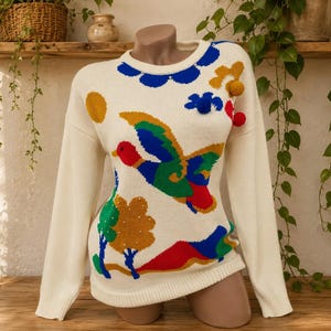May include: A cream-coloured long-sleeve knit jumper featuring a colourful intarsia design of a flying duck, trees, and a sun. The jumper includes blue scalloped trim at the neckline and three-dimensional pom-poms in yellow, blue, and red.