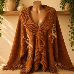 May include: A rust-coloured knit poncho featuring a wide ribbed collar, a textured surface, and fringe along the bottom hem. The garment is decorated with two symmetrical floral-shaped appliqués made of gold-toned fabric and clear crystals.