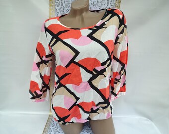 Puede incluir: Una blusa de manga larga con cuello redondo y un colorido estampado geométrico. El diseño incluye formas en rojo, rosa, beige y negro sobre un fondo blanco. Las mangas son de tres cuartos.