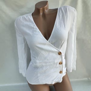 Può includere: Blusa bianca a maniche lunghe con scollo a V e tre bottoni marroni.