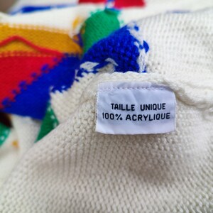May include: A cream-coloured knit sweater featuring bold, abstract geometric shapes in primary colours including red, yellow, green, and blue. A white fabric tag is sewn onto the garment, displaying the text TAILLE UNIQUE 100% ACRYLIQUE.