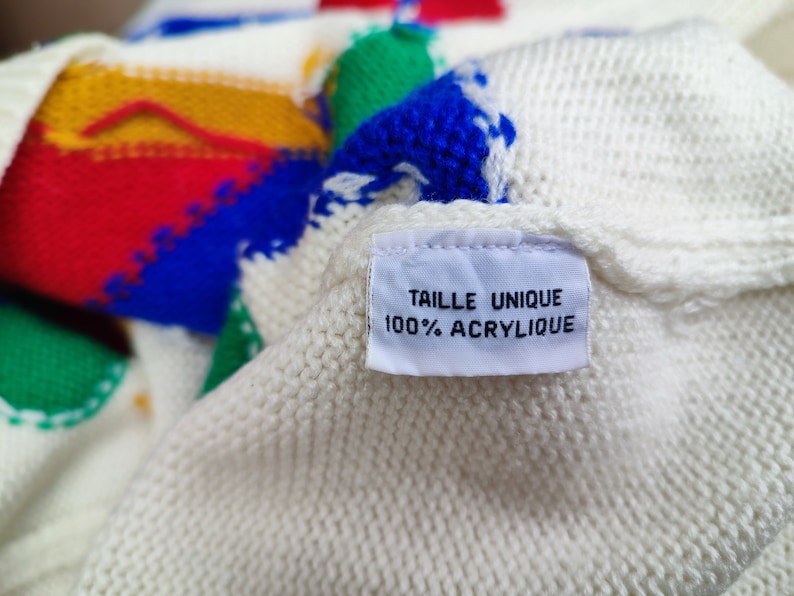 May include: A cream-coloured knit sweater featuring bold, abstract geometric shapes in primary colours including red, yellow, green, and blue. A white fabric tag is sewn onto the garment, displaying the text TAILLE UNIQUE 100% ACRYLIQUE.