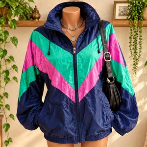 Peut inclure: Un coupe-vent bleu marine avec un motif chevron vif en rose et turquoise sur la poitrine et les manches. La veste présente un col montant, une fermeture éclair intégrale sur le devant, des manches longues à poignets élastiques et un ourlet.