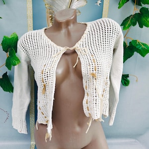 May include: Off-white, open-front crochet cardigan with long sleeves. The cardigan features a V-neck and is tied with tan-colored ties. The crochet design has a net-like appearance, with tan-colored accents throughout the garment.