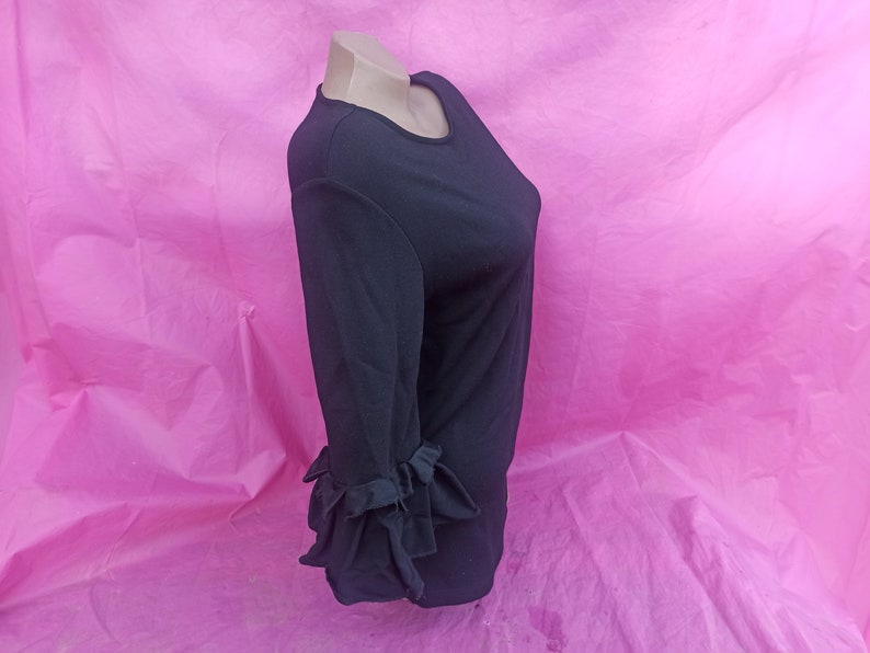 May include: Black long-sleeve shirt with ruffled cuffs. The shirt is on a pink background.
