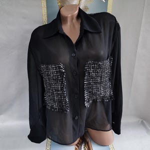 Può includere: Camicia nera trasparente con bottoni e due tasche quadrate in tweed. La camicia ha un colletto classico e maniche lunghe. Le tasche in tweed sono nere e bianche. La camicia è esposta su un manichino.