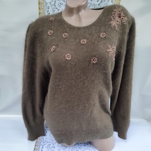 May include: A brown, long-sleeved sweater with a round neckline. The sweater is decorated with small, flower-shaped embellishments and a larger floral design on the shoulder. The fabric appears soft and fuzzy.