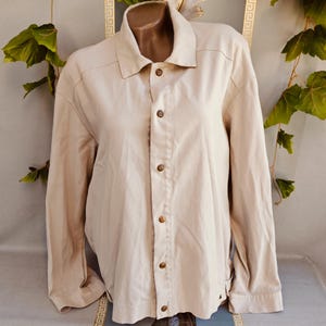 May include: A long-sleeved beige button-down shirt featuring a classic collar, a front placket with five visible metallic buttons, and structured shoulder seams. The garment is crafted from a solid-coloured fabric with a smooth, matte finish.