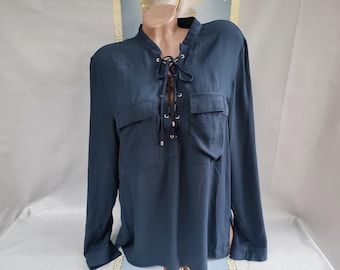 Puede incluir: Blusa de manga larga azul verdoso oscuro con escote con cordones y dos bolsillos delanteros. La blusa tiene cuello mandarín y detalles dorados en los lazos. La tela parece ser un material ligero y fluido.
