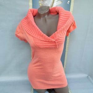 May include: Coral-colored short-sleeved sweater with a wide, ribbed cowl neck. The sweater has a fitted silhouette and is displayed on a mannequin. The neckline features a textured, ribbed design, and the body of the sweater is smooth.