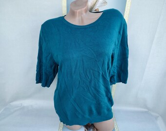 Puede incluir: Suéter de color turquesa con cuello redondo y mangas cortas. La parte superior de punto se muestra en un maniquí. La tela parece ser un material suave y ligero. El suéter es de color liso y tiene un diseño simple y clásico.
