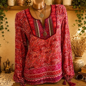 Op de afbeelding: Een blouse met lange mouwen in rood- en rozetinten met een complex paisley-patroon. Het kledingstuk heeft een split-halslijn met decoratief borduurwerk in groen, wit en donkerrood, en is voorzien van strikmanchetten met kleine kwastjes.