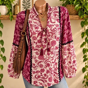 May include: A long-sleeved blouse featuring a magenta and white floral paisley pattern, a V-neckline with tassel ties, and black scalloped trim accents along the front panels and sleeves, paired with a brown leather shoulder bag with a tassel.