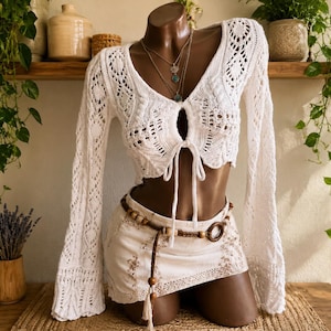 May include: A white long-sleeved crochet top with a front tie closure and a V-neckline, paired with a short white skirt featuring light brown floral embroidery and a brown braided belt with wooden beads and a circular wooden buckle.