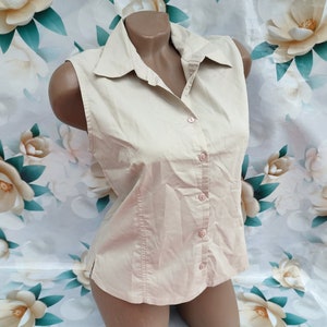 Può includere: Una camicia senza maniche di colore beige chiaro con bottoni. La camicia ha un colletto e una chiusura con bottoni sul davanti.