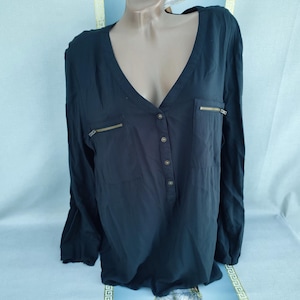 May include: Black long-sleeved blouse with a V-neck and two chest pockets. The blouse has gold-coloured zips on the pockets and a button-down front. The fabric appears to be a lightweight material.