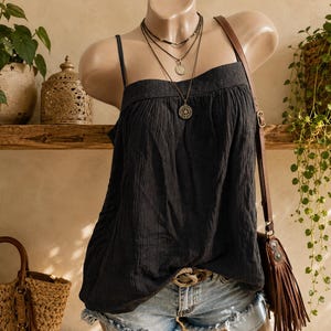 May include: A black sleeveless top with a textured fabric finish, worn with a layered necklace featuring three metallic pendants. The top is paired with light blue denim shorts and a brown leather crossbody bag with fringe detailing on the side.