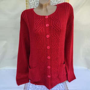 May include: A red knitted cardigan with a cable knit pattern and five pink buttons down the front. The cardigan has two pockets on the bottom.