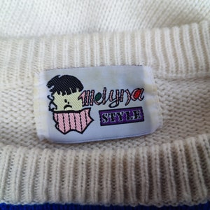 May include: A cream-coloured knit jumper featuring a rectangular fabric label at the inner neckline. The label displays a graphic of a person with dark hair and a pink scarf, alongside the text Melyna STYLE in purple and black lettering.