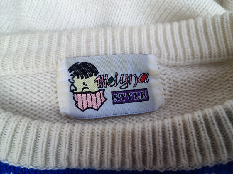 May include: A cream-coloured knit jumper featuring a rectangular fabric label at the inner neckline. The label displays a graphic of a person with dark hair and a pink scarf, alongside the text Melyna STYLE in purple and black lettering.