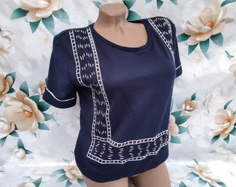 Puede incluir: Camiseta azul marino de manga corta con detalles bordados en blanco. La camiseta presenta un cuello redondo y ribetes blancos en las mangas. El diseño bordado es un patrón geométrico. La camiseta se exhibe en un maniquí.