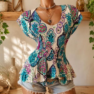 May include: A short-sleeved, pleated top featuring a vibrant, multi-colored abstract pattern in shades of teal, magenta, and dark blue on a cream background, paired with light blue denim shorts and a layered gold-tone chain necklace with a heart pendant.