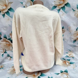 May include: A cream-coloured long-sleeve knit jumper featuring a ribbed crew neckline, cuffs, and hem. The back of the garment displays a solid, plain knit texture in a light off-white shade, with a small portion of a blue pattern visible.