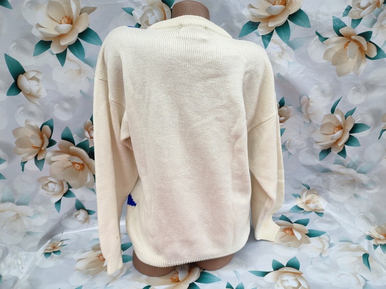 May include: A cream-coloured long-sleeve knit jumper featuring a ribbed crew neckline, cuffs, and hem. The back of the garment displays a solid, plain knit texture in a light off-white shade, with a small portion of a blue pattern visible.