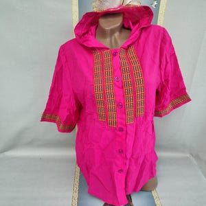 Puede incluir: Una camisa rosa brillante con botones y capucha. La camisa tiene mangas cortas y rayas verticales decorativas en naranja, negro y amarillo. Los puños y la capucha tienen patrones a juego. Una prenda colorida para cualquier guardarropa.