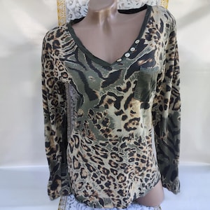 May include: Long-sleeved V-neck shirt with a leopard print design in shades of brown, black, and olive green. Features a small pocket and three white buttons. The shirt is displayed on a mannequin.