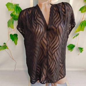 May include: A black semi-sheer blouse featuring a V-neckline and short sleeves. The fabric is decorated with a repeating vertical wave pattern created by textured, opaque black lines that contrast against the translucent sections of the garment.