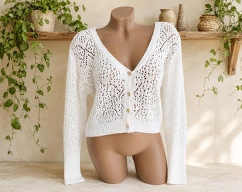 May include: A white long-sleeve cardigan featuring an open-knit crochet pattern, a deep V-neckline, and a front button closure with four light-colored buttons. The garment has ribbed cuffs and a ribbed hem, showcasing a lightweight, textured design.