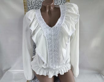 May include: A white blouse with long sleeves, a V-neck, and ruffled details. The blouse features intricate white lace trim along the neckline and down the center. The sleeves have a slight puff at the shoulders.