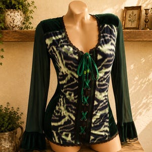 Può includere: Un top a maniche lunghe con corpetto in velluto verde scuro e un pannello centrale in tessuto con motivo astratto nero e verde chiaro. La parte anteriore presenta un'allacciatura verticale con nastro verde, scollo rotondo e polsini svasati.