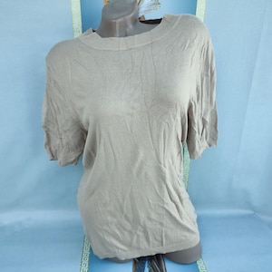 May include: A short-sleeved, light brown knit top with a crew neck. The shirt is displayed on a mannequin against a light blue background. The fabric appears soft and slightly textured.