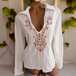 Puede incluir: Una camisa blanca de manga larga con un escote en V profundo y detalles de bordado intrincado en tonos marrón y dorado alrededor del cuello y el pecho. La camisa cuenta con un cuello clásico, mangas largas con puños y un corte entallado.