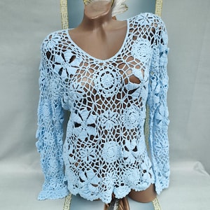 Puede incluir: Un top de ganchillo azul claro de tejido abierto, con cuello en V y mangas largas. El top presenta un patrón floral. La prenda se muestra en un maniquí.