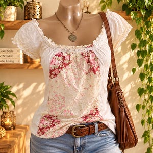 May include: A white short-sleeved blouse featuring a central panel with a pink and red floral pattern, lace trim along the scoop neckline, and eyelet fabric details. The blouse is paired with a brown leather belt and a brown leather shoulder bag.