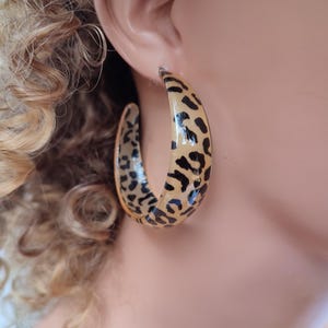 Puede incluir: Un par de pendientes grandes de aro con diseño de estampado de leopardo. Los aros son de un material marrón claro con manchas negras.
