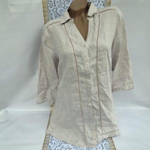 May include: A beige linen shirt with a button-up front and a delicate lace trim. The shirt has a relaxed fit and three-quarter sleeves.