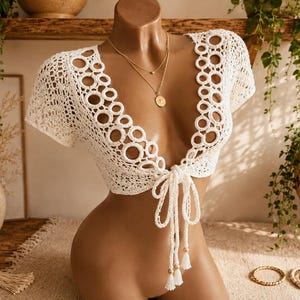 May include: A white crochet crop top featuring short sleeves, a deep V-neckline, and a front tie closure. The garment is detailed with a series of circular ring patterns along the neckline and edges, finished with tassels at the tie ends.