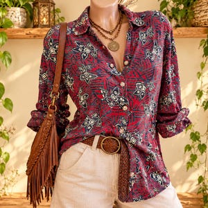 May include: A long-sleeved button-down shirt featuring a dark red, navy blue, and white geometric pattern. The shirt is paired with light beige trousers, a brown braided leather belt, and a brown leather fringe shoulder bag with a gold clasp.