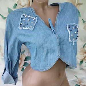 Peut inclure: Une veste en jean bleu clair à col en V et manches longues. La veste est courte et ornée de perles et de perles bleues sur le devant.