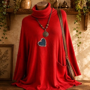 May include: A bright red turtleneck sweater featuring a blue denim heart-shaped patch on the chest, long sleeves, and a side pocket. The sweater is styled with a dark metal chain necklace with a round pendant and a brown leather shoulder bag.